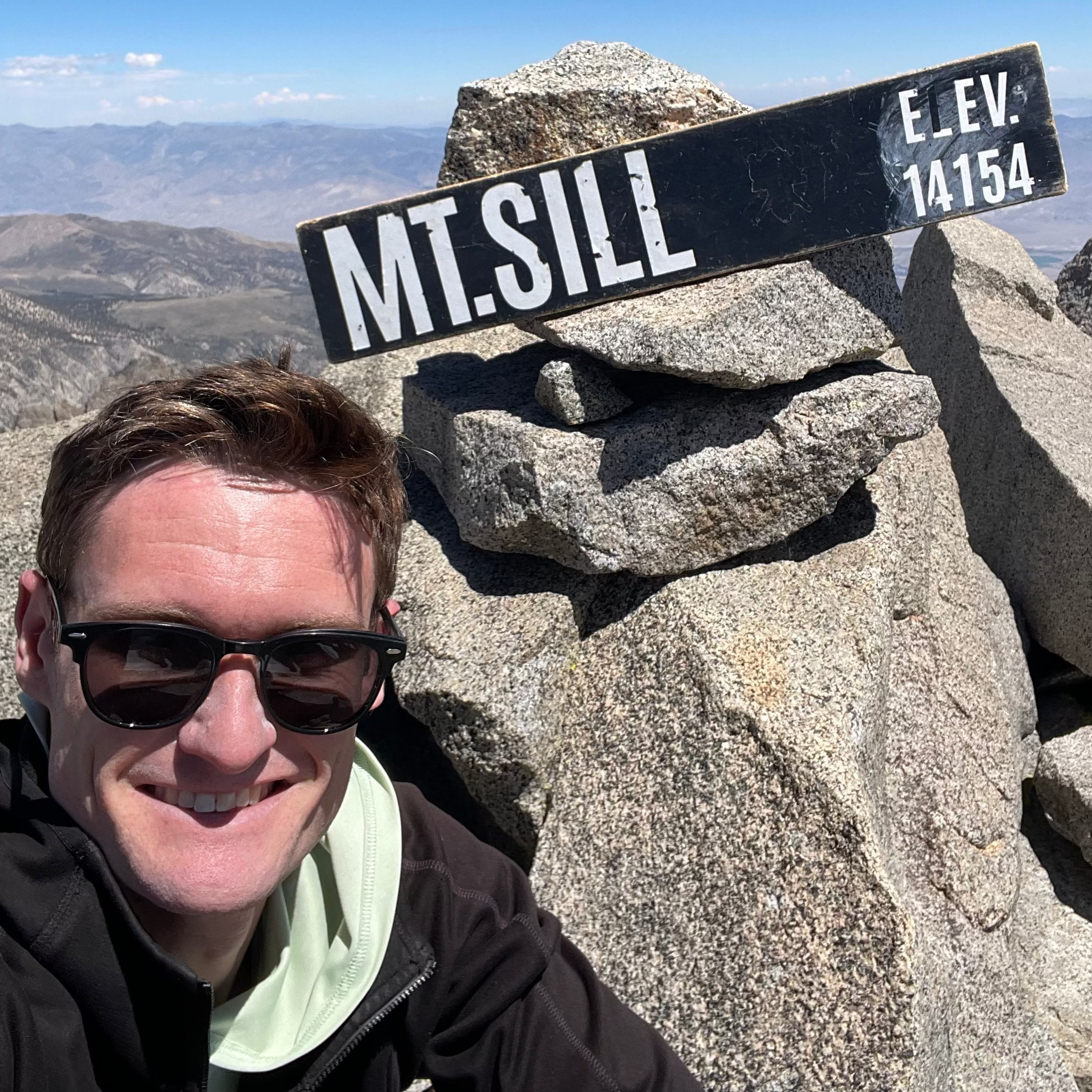 A man wearing sunglasses takes a selfie next to a black 'MT. SILL' elevation sign on a rocky mountain peak under a clear blue sky.