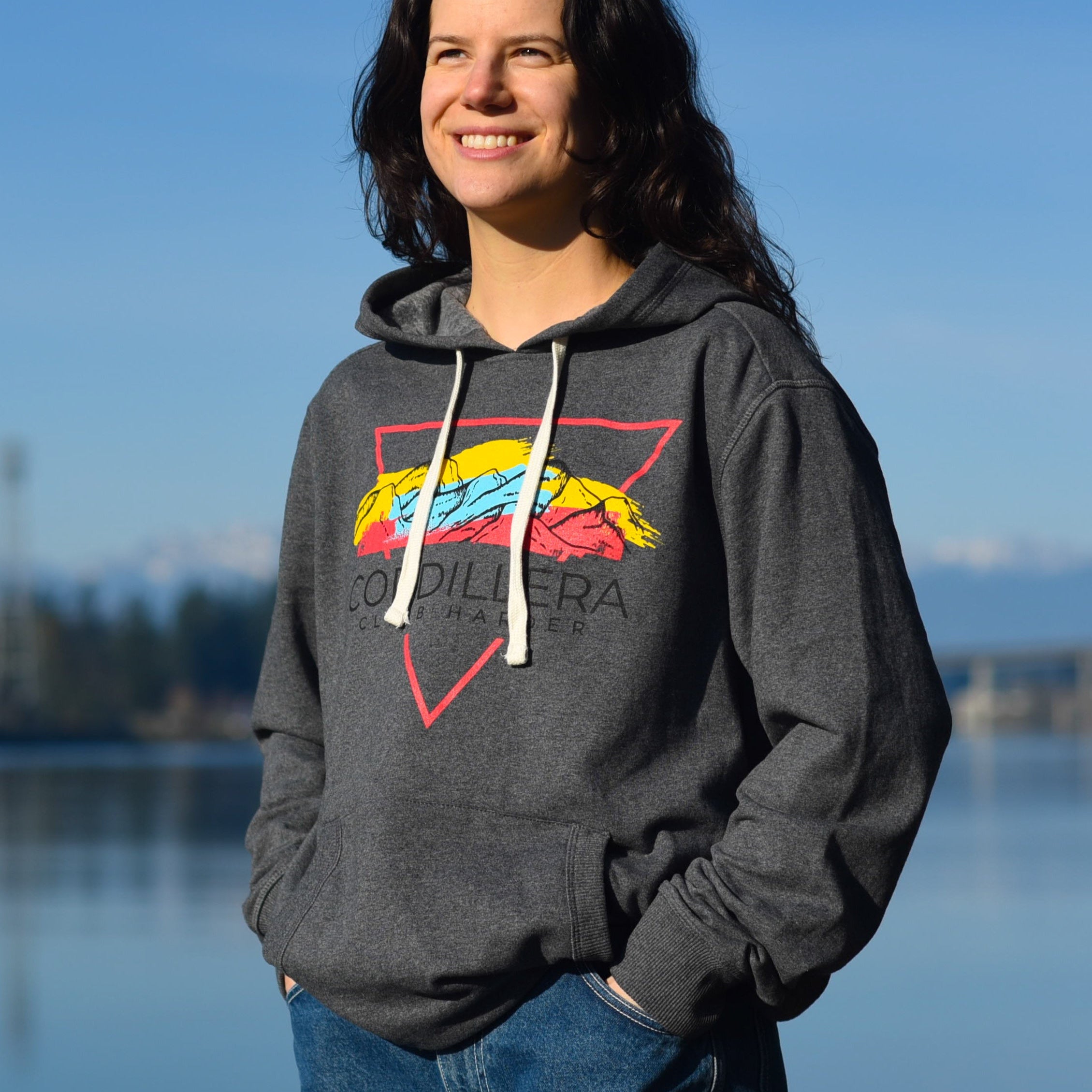Person wearing a gray hoodie with a colorful logo by a body of water